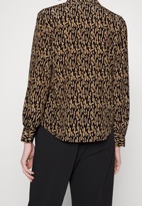 Person wearing a brown and black geometric patterned long-sleeve blouse and black pants, shown from the back with left hand relaxed.