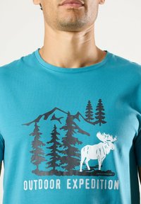 Türkises Baumwoll-T-Shirt mit einer schwarzen Grafik von Bergen und Bäumen, darunter ein weißer Elch und der Text "OUTDOOR EXPEDITION".