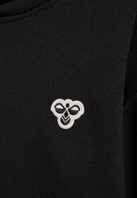 Czarny dzianinowy materiał z małym, białym haftowanym logo przypominającym pszczołę, z prostymi, stylizowanymi skrzydłami i ciałem na lewej stronie klatki piersiowej.