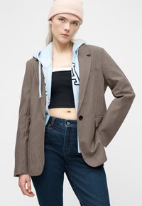 Νεαρή γυναίκα με μπεζ σκουφάκι, γαλάζιο φούτερ με κουκούλα κάτω από καφέ καρό σακάκι, μαύρο crop top και ψηλόμεσο σκούρο μπλε τζιν.