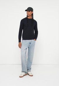 Pulover tricotat cu nervuri de culoare bleumarin, blugi largi de culoare albastru deschis și adidași deschiși. O șapcă neagră completează ținuta casual. Fundal minimalist.