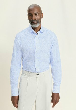 Mann mit grauem Bart, der ein hellblau gemustertes langärmliges Hemd und weiße Hose trägt, vor einem einfarbigen Hintergrund steht.