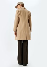 Abrigo de lana beige con un diseño entallado, detalle de pliegue en la parte posterior y puños abotonados. Combinado con pantalones oscuros de pierna ancha y una gorra de cuadros.