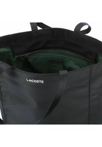 Zwarte nylon draagtas met een groene binnenzijde, voorzien van een bovenste ritssluiting en een merklogo aan de buitenkant. Robuuste stoffen handvatten inbegrepen.