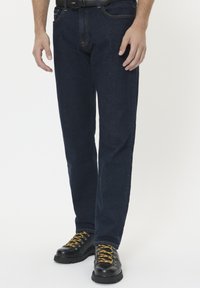 Uomo con jeans blu scuro, stivali di pelle nera con lacci gialli e una cintura nera su uno sfondo bianco.