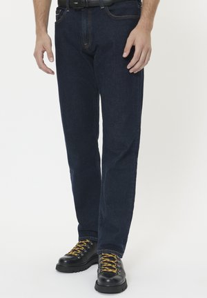 Bărbat îmbrăcat cu blugi de culoare albastru închis, cizme din piele negre cu șireturi galbene și o curea neagră pe un fundal alb.