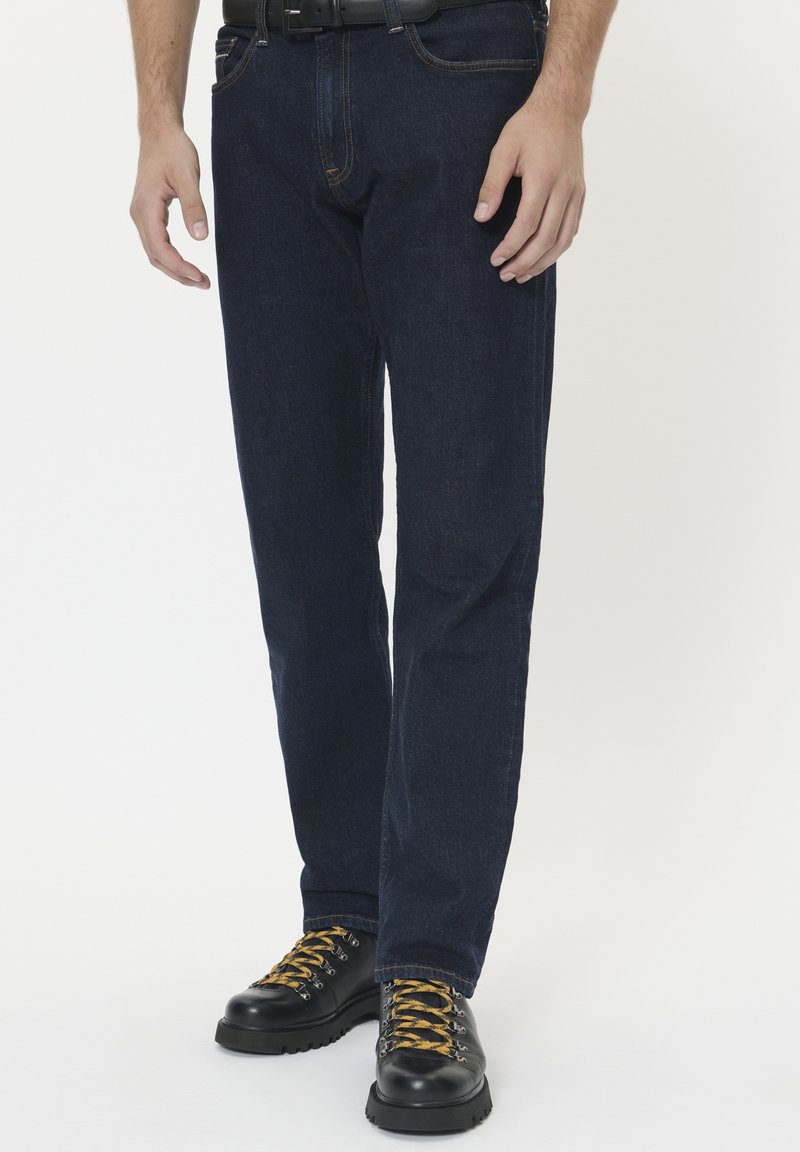 Uomo con jeans blu scuro, stivali di pelle nera con lacci gialli e una cintura nera su uno sfondo bianco.