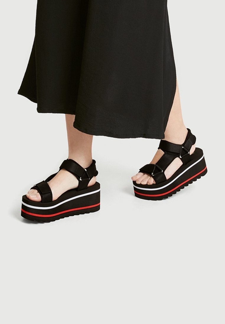 Sandales à plateforme noires avec semelles épaisses, rayures blanches et rouges, sangles réglables et surface texturée. Portées avec une jupe noire.