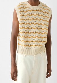 Ärmlös beige stickad tröja med vågiga orange ränder, bärs över vita byxor med hög midja, visad från halsen till höfterna.