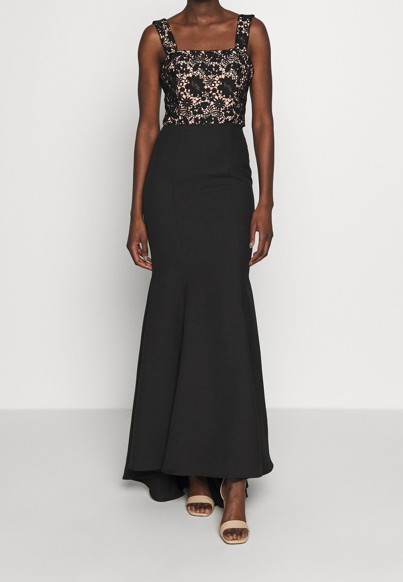 Femme portant une robe noire sans manches avec un corsage en dentelle florale et une longue jupe ajustée, associée à des sandales beiges à bouts ouverts.