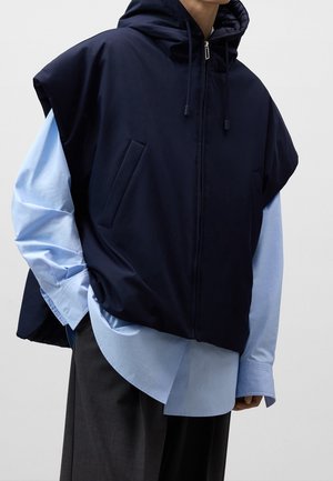 Person iført navy ærmeløs hættetrøje over lyseblå langærmet skjorte med sorte bukser, hånd i vestlommen, stående mod en ensfarvet baggrund.