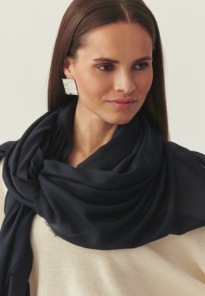 Nő hosszú barna hajjal, nagy, texturált ezüst fülbevalóval, fekete sállal a nyakában és bézs kötött pulóverben.