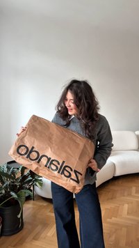 Hosszú, sötét hajú nő egy nagy barna papír Zalando bevásárlószatyorral a fehér kanapé mellett, zöld cserépben lévő növény közelében.