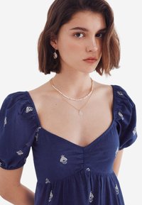 Tummansininen puff-hihainen mekko, jossa on valkoista kirjontaa, syvä V-muotoinen kaula-aukko ja rypytetty vyötärö. Kerrokselliset helmi- ja kultaketjut.