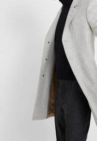 Abrigo de lana gris claro con diseño entallado, cierre de un solo botón e interior negro, llevado sobre un suéter de cuello alto negro acanalado y pantalones oscuros.