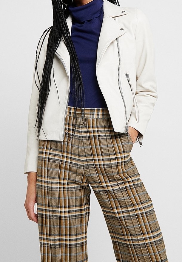 Bílá kožená bikerská bunda se zipy, nošená přes modrý rolák a kárované široké kalhoty v odstínech hnědé a žluté.