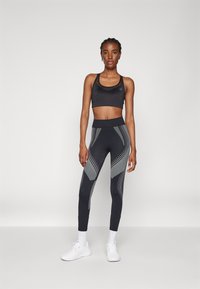 Sutien sport negru cu bretele late și leggings negri cu talie înaltă, prezentând modele geometrice gri și accente punctate. Pantofi sport albi.