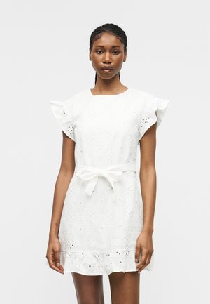 Žena stojí v bielych šatách s výšivkou, s priesvitnými rukávmi, s mašľou v páse a s volánovým lemom na jednoduchom svetlom pozadí.