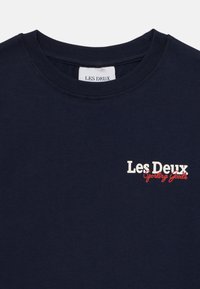 T-shirt de algodão azul-marinho com gola redonda. Apresenta um logótipo gráfico "Les Deux Sporting Goods" em branco e vermelho no peito.