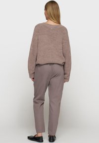 Kobieta z długimi blond włosami, ubrana w fioletowy sweter z miękkiego materiału, dopasowane fioletowe spodnie i czarne loafersy, stojąca plecami na białym tle.