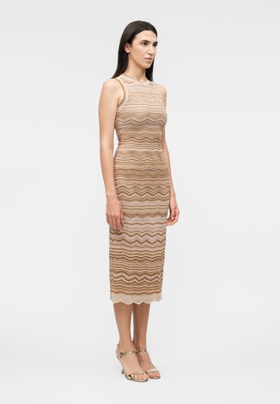 Γυναίκα με μακριά σκούρα μαλλιά στέκεται φορώντας ένα αμάνικο, μέχρι το γόνατο, μπεζ και καφέ φόρεμα με κυματιστό μοτίβο και χρυσά ανοιχτά πέδιλα.