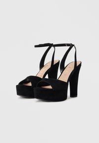 Zwarte plateauhoge sandalen met enkelbandjes en open tenen, met de merknaam "GUESS" op de beige voetbedden.