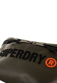 Oliwkowa nylonowa saszetka z czarnym haftowanym napisem "SUPERDRY" oraz pomarańczowym logo. Posiada zamek błyskawiczny z teksturowanymi uchwytami.