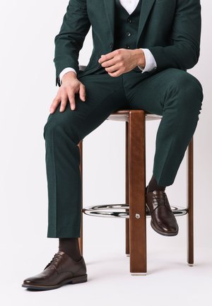 Man in dark green suit and brown dress shoes sitting on a wooden stool with metal footrest against a white background.