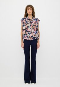 Top senza maniche con motivo floreale in tessuto morbido e collo drappeggiato. Indossato con pantaloni a zampa neri. Il top presenta colori pastello su uno sfondo navy.