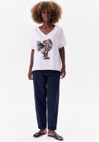 Witte oversized t-shirt met een kleurrafische afbeelding van een vrouw met een gemusterd hoofddeksel, gecombineerd met donkerblauwe broek en zwarte slippers.