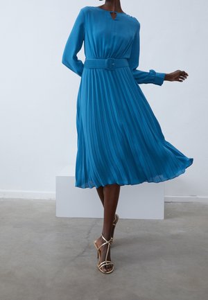 Femme portant une robe plissée bleue à manches longues avec ceinture, associée à des sandales à talons hauts beige, en mouvement sur un sol en béton.
