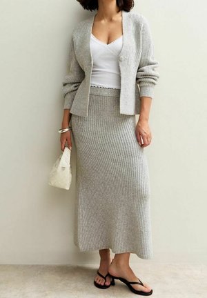 Femme portant un ensemble cardigan tricoté gris et jupe, camisole blanche, sandales à talons noires, tenant un petit sac à main blanc tissé.