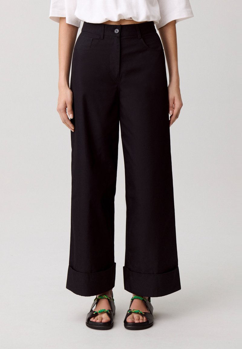 Pantaloni neri a gamba larga con chiusura a bottone, tasche frontali e orli risvoltati. Il tessuto appare liscio con una sartoria strutturata.