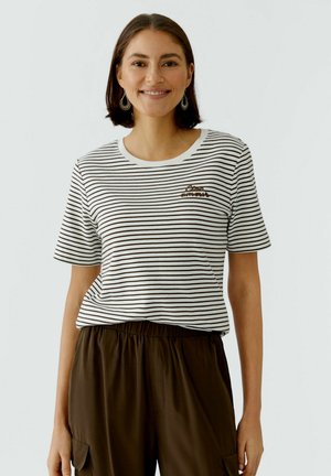 Femme aux cheveux bruns courts souriant, portant un t-shirt rayé noir et blanc avec le texte « Ciao amore » et un pantalon large marron foncé.