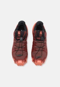 Încălțăminte sportivă roșie și neagră cu un upper texturat, închidere cu șireturi și talpă robustă; prezintă un logo proeminent și un design solid.