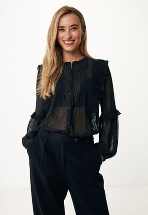 Blusa nera trasparente con maniche arricciate e pieghe verticali. Presenta una chiusura con bottoni e un piccolo motivo a pois, abbinata a pantaloni a vita alta.