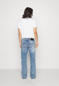 Una persona que lleva una camisa blanca y jeans acampanados azules, mostrando un denim de lavado claro, con bolsillos traseros con detalles de costura y un objeto negro en la cintura.