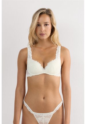 Kobieta nosząca biały koronkowy biustonosz i pasujące koronkowe majtki, stojąca na jasnym, jednolitym tle, zwrócona przodem.