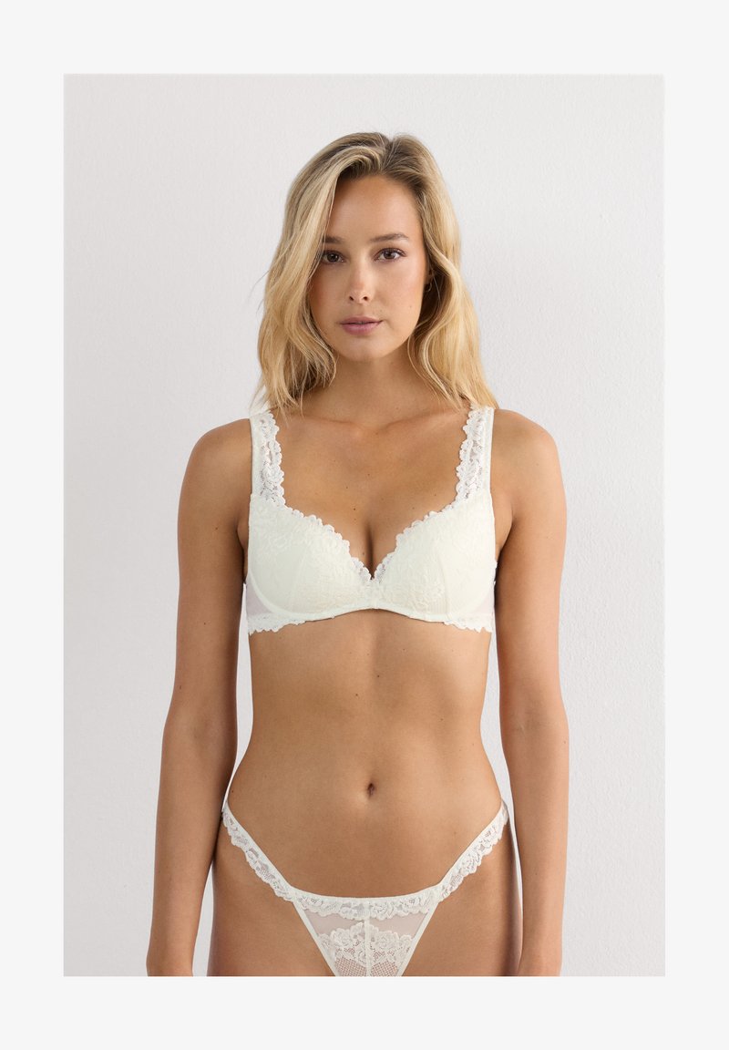 Kvinde iført en hvid blonde-bh og matchende blonde-trusser, stående mod en lys ensfarvet baggrund, vendt mod kameraet.