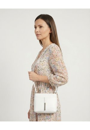 Kvinde i en pastelblomstret kjole, der bærer en lille hvid crossbody-taske med en metalspænde foran, stående mod en ensfarvet lys baggrund.