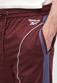 Bordó sport nadrág elasztikus derékpánttal, húzózsinórral, fehér díszítéssel és oldalán zsebekkel, kék részletekkel. Reebok logóval.