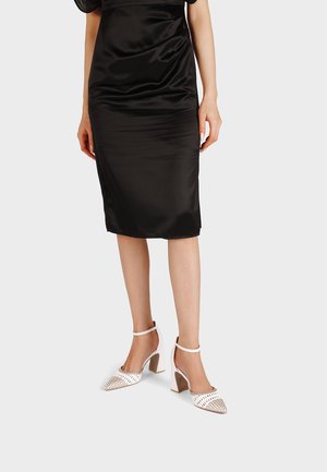 Donna con gonna nera in raso fino al ginocchio e tacchi alti bianchi con cinturino alla caviglia e punte appuntite borchiate su sfondo chiaro.