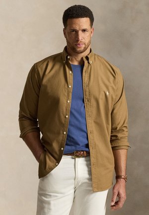 Lühikeste lokkis juustega mees, kes kannab sinist särki lahtise pruuni nööbitava särke ja valkjaid pükse taustal, mis on neutraalne.