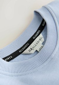 Šviesiai mėlynos spalvos džemperio apykaklė su ribbingu, ant kurio yra juoda etikėtė su "AllSaints" prekės ženklu ir priežiūros informacija po ja.