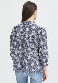Blouse bleu marine à motifs floraux blancs en dentelle, manches bouffantes et ourlet festonné. Confectionnée dans un tissu léger pour une apparence texturée.