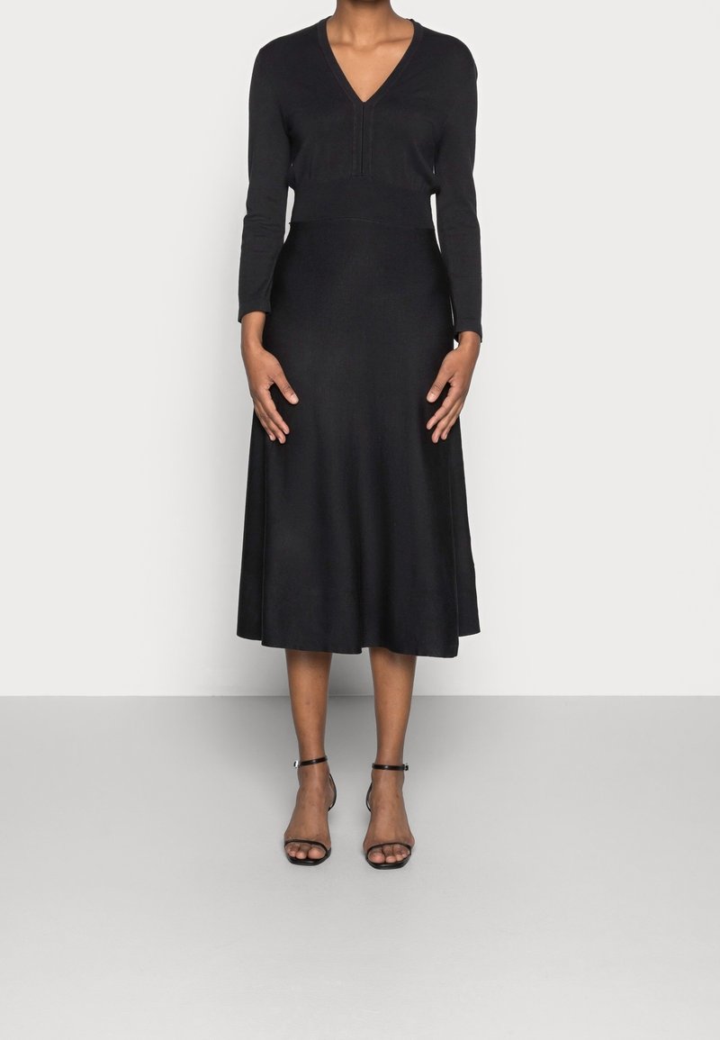Svart långärmad klänning med V-ringning, figursydd överdel och utsvängd kjol. Slät textur, knä-lång, matchad med svarta sandaletter med remmar.