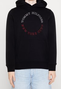 Svart hoodie med framficka, med vitt och rött cirkulärt text "TOMMY HILFIGER NEW YORK CITY." Mjukt, strukturerat tyg, avslappnad passform.