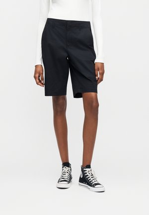 Άτομο που φοράει μαύρο σορτς μέχρι το γόνατο, λευκό μακρυμάνικο μπλουζάκι και μαύρα παπούτσια τύπου high-top, στέκεται μπροστά σε απλό φόντο.