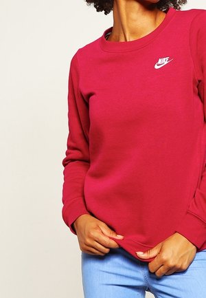 Persoon die een rood Nike sweatshirt draagt met een wit logo op de borst, gecombineerd met lichtblauwe jeans, tegen een effen lichte achtergrond.