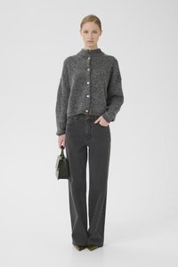 Grijze gebreide cardigan met een hoge kraag en knopen aan de voorkant, gecombineerd met hoog-gesneden zwarte jeans. Model houdt een kleine zwarte handtas vast.